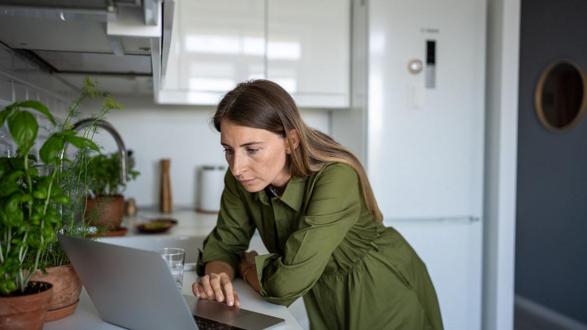 Eine Frau in einem grünen Kleid steht in einer Küche und schaut kritisch auf ihren Laptop