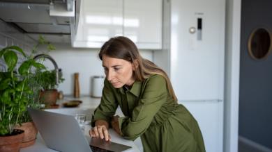 Eine Frau in einem grünen Kleid steht in einer Küche und schaut kritisch auf ihren Laptop