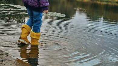 Kind in lilafarbener Jacke mit gelben Gummistiefeln in einer Pfütze
