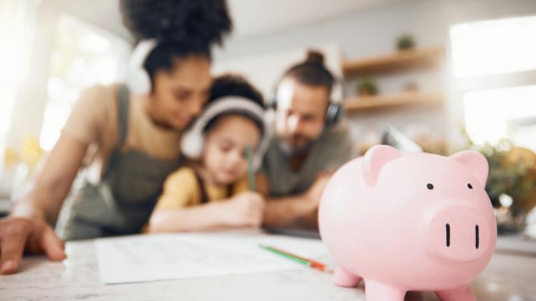 Familie sitzt am Tisch. Im Vordergrund steht ein Sparschwein 
