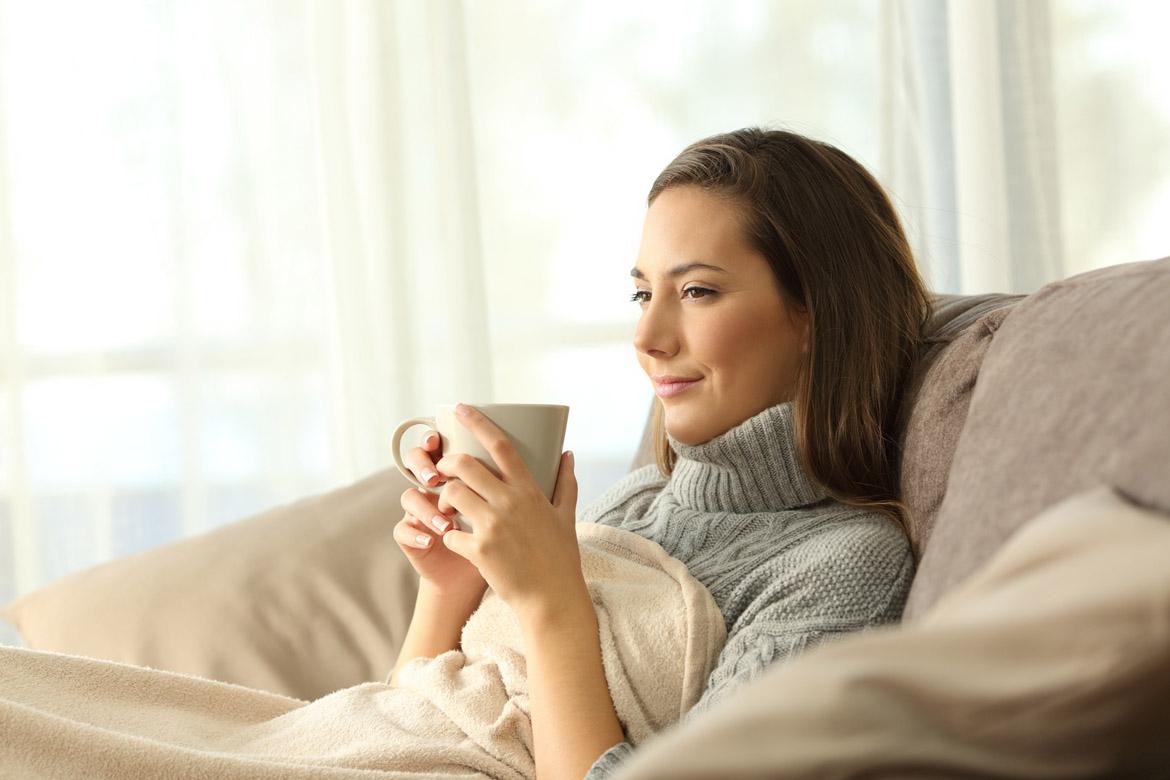 Frau sitzt mit einer Tasse und Decke auf dem Sofa