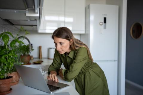 Eine Frau in einem grünen Kleid steht in einer Küche und schaut kritisch auf ihren Laptop