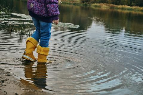 Kind in lilafarbener Jacke mit gelben Gummistiefeln in einer Pfütze