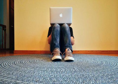 Junger Mensch hat Laptop auf Knien und sitzt auf dem Fußboden