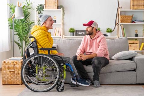 Mann mit körperlicher Behinderung im Rollstuhl und Betreuer, der mit ihm entspannt zu Hause spricht