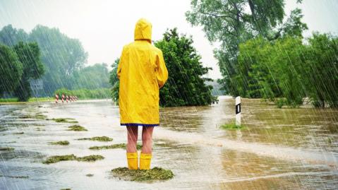 Frau steht im Regenmantel und Gummistiefeln auf überschwemmter Straße 